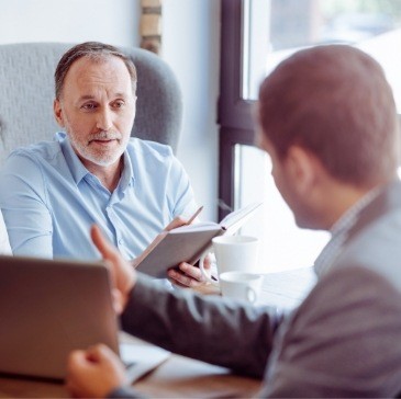 Man with notebook talking to man with laptop from across desk