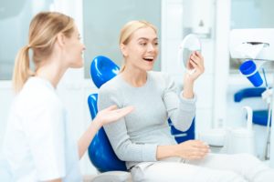 Blond woman in gray sweater sitting in blue dental chair holding a mirror smiling at her reflection with a dentist sitting next to her