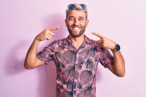 Man in a pink shirt and sunglasses pointing to his smile. 